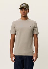 Γκρι βαμβακερό μπλουζάκι με κοντά μανίκια, στρογγυλή λαιμόκοψη και μικρό σκούρο λογότυπο στο στήθος. Συνδυασμένο με μπλε τζιν παντελόνι και πράσινο καπέλο.