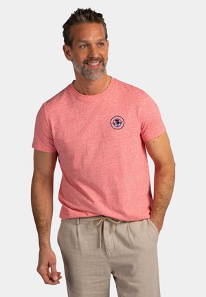 Man met baard die een roze T-shirt met korte mouwen en ronde hals draagt met een klein embleem en lichtbeige broek met trekkoord, glimlachend en poseerend tegen een effen achtergrond.