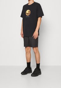 Homme portant un T-shirt noir avec un grand graphique de lune jaune, un short à carreaux sombre, des chaussettes noires et des baskets noires, se tenant devant un fond blanc.