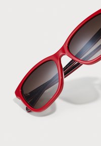 Red sunglasses with large, angular frames and dark gradient lenses. The arms have a glossy finish with subtle markings.