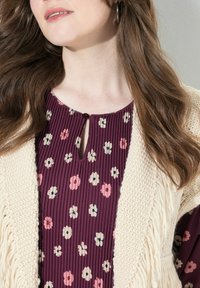 Pflaumenfarbene Bluse mit floralem Muster in Pink und Creme, ausgestattet mit einem Schlüsselloch-Ausschnitt; kombiniert mit einer cremefarbenen Strickjacke mit Fransen.