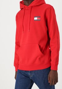 Felpa rossa in cotone, con tasca frontale e cappuccio regolabile con lacci. Include una patch con logo in blu e bianco.