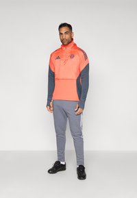 Ανδρικό αθλητικό σύνολο που περιλαμβάνει μισό φερμουάρ σε φωτεινό πορτοκαλί και σκούρο μπλε χρώμα με λογότυπο, σε συνδυασμό με γκρι φόρμες και μαύρα παπούτσια.