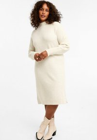 Cremfarbener Strickkleid mit hohem Kragen, gerippten Manschetten und gerader Silhouette, mit seitlichen Schlitzen für Bewegungsfreiheit.