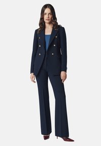 Granatowy, dobrze skrojony garnitur z podwójnym zapięciem, ozdobiony złotymi guzikami, w zestawieniu z rozszerzanymi spodniami i dopasowaną niebieską bluzką.