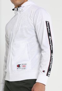 Vit huvtröja med huva som har dragkedja, mönstrad logotyptryck och en märkesstripe på ärmen med röda och svarta detaljer.