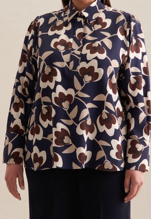 Blůza s dlouhým rukávem s námořnicky modrým, béžovým a hnědým květinovým vzorem, se špičatým límcem a zapínáním na knoflíky na manžetách, nošená s tmavými kalhotami.