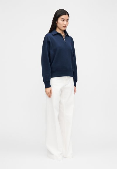 Tumesinine dressipluus lukuga krae ja laiade varrukatega, kombineeritud valgete laiade pükstega. Lihtsad, ühtlased värvid minimaalse detailiga.