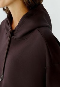 Übergroßer brauner Hoodie mit einer Kordelzug-Halslinie, glatter Textur und Seitentasche, die ein entspanntes, lässiges Design betont.