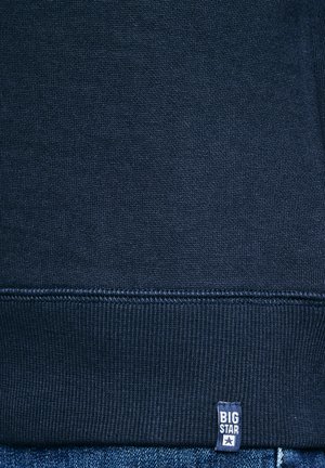 Material tricot albastru marin cu tiv răsfrânt și o etichetă dreptunghiulară mică pe marginea de jos, pe care scrie "BIG STAR" și are o iconiță în formă de stea.