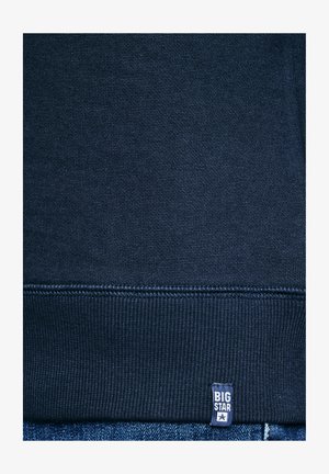 Tengerészkék kötött anyag bordázott szegéllyel és egy kis téglalap alakú címkével, amelyen „BIG STAR” felirat és egy csillag ikon található az alsó szél közelében.