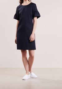 Vestido azul marino con mangas cortas con volantes, corte recto y acentos de rayas laterales. Usado con zapatos blancos de tipo slip-on. Textura de tejido suave.