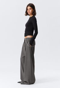 Femme debout de profil portant un haut noir ajusté à manches longues, un pantalon large à rayures grises avec des détails en chaîne et des chaussures noires à talons pointus.