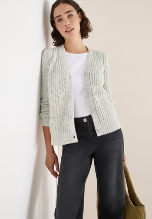 Helle grüne Strickjacke mit gerippter Textur, Knopfverschluss und zwei vorderen Taschen, getragen über einem weißen Oberteil und dunklen Jeans.