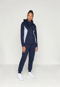 Ναυτικό αθλητικό σετ με φούτερ με φερμουάρ που διαθέτει ανοιχτό γκρι λεπτομέρειες και λογότυπο. Το παντελόνι είναι σε στενή γραμμή με ελαστικές μανσέτες.