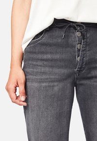 Jeans de mezclilla gris oscuro con un diseño ajustado, que cuentan con un cordón y un cierre de botón visible, mostrados con una mano descansando sobre el lado.