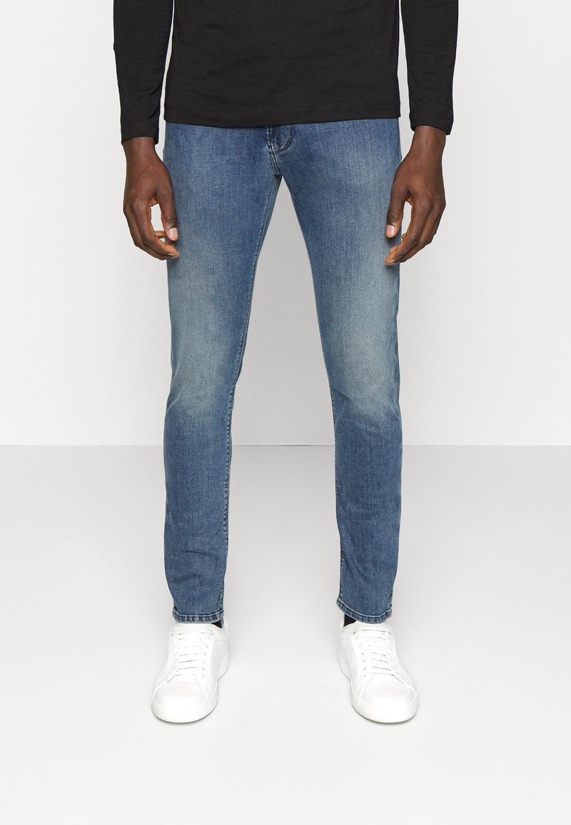Blå denimjeans med slim fit, med en bleknad tvätt, femficksdesign och avslutad med vita sneakers och en svart långärmad skjorta.