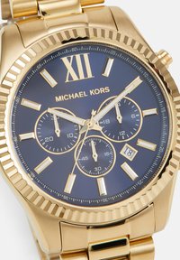 Goldene Michael Kors Armbanduhr mit blauem Zifferblatt, römischer Zahl XII, drei Hilfszifferblättern, Datumsanzeige auf 18 und goldenen Stundenmarkierungen und Zeigern.