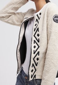 Kurtka polarowa w kolorze kremowym z czarnym geometrycznym wzorem wzdłuż zamka. Charakteryzuje się miękką fakturą i prążkowanymi krawędziami; zapięcie na zamek błyskawiczny z przodu.
