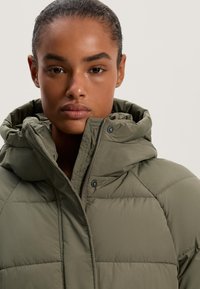 Olive zöld puffernadrág magas gallérral és kapucnival, steppelt dizájnnal, elülső cipzárral és snap gombokkal. Simább, matt textúra.