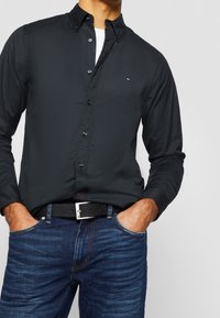 Musta napillinen paita, jossa on jämäkkä kaulus, kapea leikkaus ja hillitty logo. Yhdistetty sinisiin farkkuihin ja mustaan vyöhön, jossa on hopeinen solki.