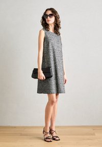 Grijze mouwloze jurk met een textuurpatroon, gecombineerd met een zwarte clutch en zwarte sandaaltjes met bandjes, gedragen door een model tegen een neutrale achtergrond.