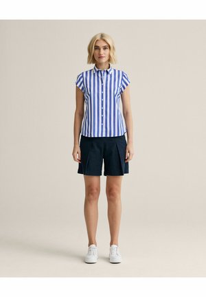 Kvinde stående iført en blå og hvid kortærmet skjorte med lodrette striber, marineblå shorts og hvide sneakers på beige baggrund.