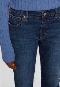 Jeans de denim en azul oscuro con una textura fina, diseño de cinco bolsillos, costuras naranjas y cierre de botón de metal. Llevado con un suéter de punto azul claro.