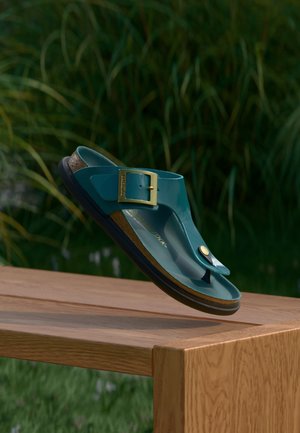 Sandale bride teal avec boucle dorée et semelle en liège flottant au-dessus d'un banc en bois avec des plantes vertes floues en arrière-plan.