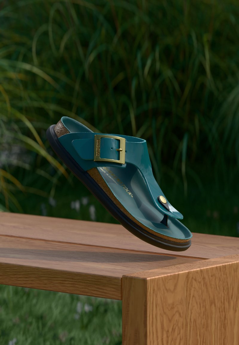 Σανδάλι τύπου thong σε τυρκουάζ με χρυσή αγκράφα και φελλό σόλα που αιωρείται πάνω από ένα ξύλινο παγκάκι με θολά πράσινα φυτά στο φόντο.