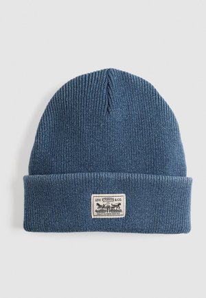 Blue knit beanie hat with a folded cuff, featuring a sewn-on label with a logo and text. The texture is ribbed and made from soft material.