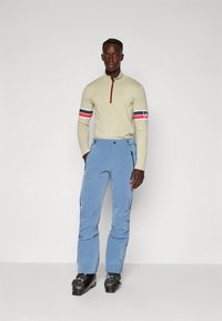 Beige langermet topp med kvartglidelås og rødt aksent. Lyseblå bukser med lommer. Sorte skistøvler med justerbare spenner.