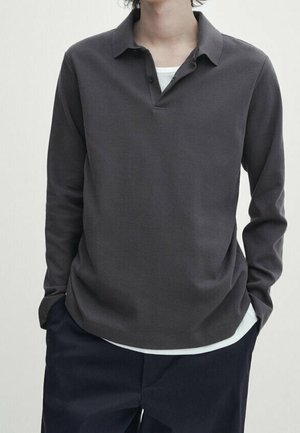 Osoba ubrana w ciemnoszary długi rękaw polo na białej podkoszulce i ciemnogranatowe spodnie, z rękami w kieszeniach, na jednolitym tle.