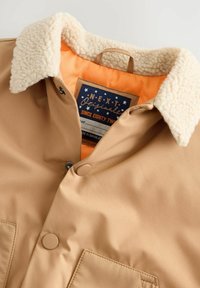 Chaqueta marrón claro con un suave collar de sherpa beige, forro naranja, dos bolsillos frontales y botones a presión. La etiqueta presenta estrellas en color marino y blanco.