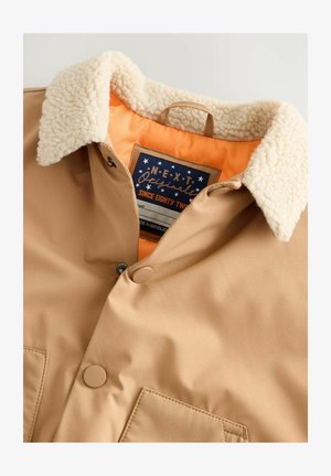 Barna dzseki puha, bézs sherpa gallérral, narancssárga béléssel, két elülső zsebbel és patent gombokkal. A címke sötétkék és fehér csillagokat tartalmaz.