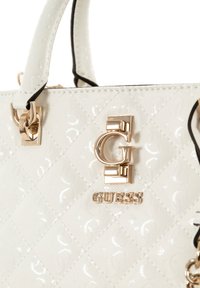 Bolso acolchado blanco hecho de material brillante con herrajes dorados. Presenta un logotipo prominente y costuras de acento negro.