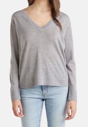 Femme portant un pull gris clair à col en V et un jean bleu clair déchiré, debout devant un fond blanc uni.