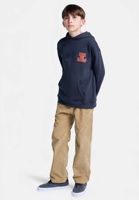 Navy hoodie with orange "Ride the Rails" text, kangaroo pocket, paired with tan corduroy pants and gray sneakers. Simple design, casual fit.