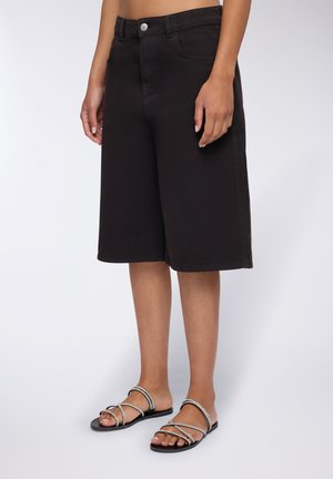 Culotte in denim nero con taglio dritto, dotate di tasche frontali e un singolo bottone argentato. Abbinato a sandali neri con cinturini.