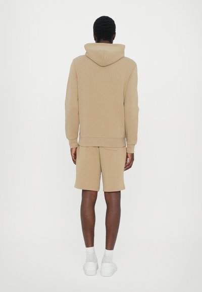 Beige hoodie and shorts set made of soft fabric. The hoodie features a drawstring hood. Model wears white socks and chunky white shoes.