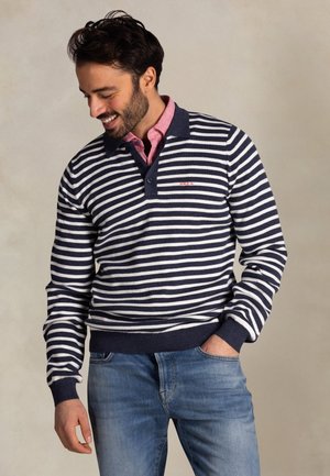 Man lacht, draagt een marineblauw-wit gestreepte polo met lange mouwen over een roze shirt en een blauwe spijkerbroek, en staat voor een effen beige achtergrond.