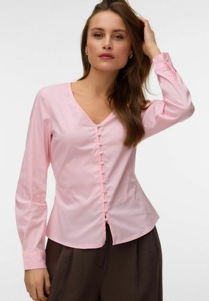 Žena v svetloružovej blúzke s dlhým rukávom a prednými gombíkmi pózuje so zdvihnutou rukou, ktorá sa dotýka jej vlasov, a nosí tmavé nohavice.