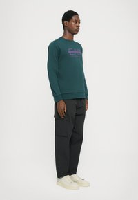 Sweatshirt verde com logótipo roxo, combinado com calças cargo pretas com bolsos e sapatilhas em beige claro. Tecido suave, ajuste casual.