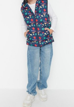 Kind draagt een navyblauw gewatteerd vest met kleurrijke geometrische vormen, een wit shirt met lange mouwen, losse blauwe spijkerbroek en witte sneakers.