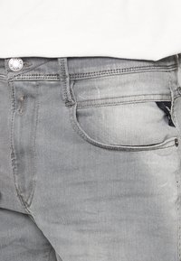 Jeans de mezclilla gris con un acabado desgastado, cinco bolsillos y botón de metal. La tela tiene una textura suave y un ligero estiramiento para mayor comodidad.