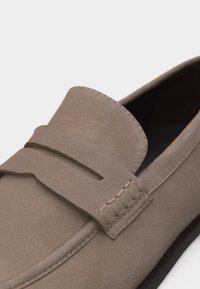 Mocassin en cuir suédé taupe avec des détails cousus et une bride classique à lanière sur le dessus, présenté en vue rapprochée et en angle.