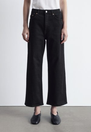 Person wearing high-waist, wide-leg black jeans with five pockets and black slip-on shoes, standing on a gray floor against a white wall.