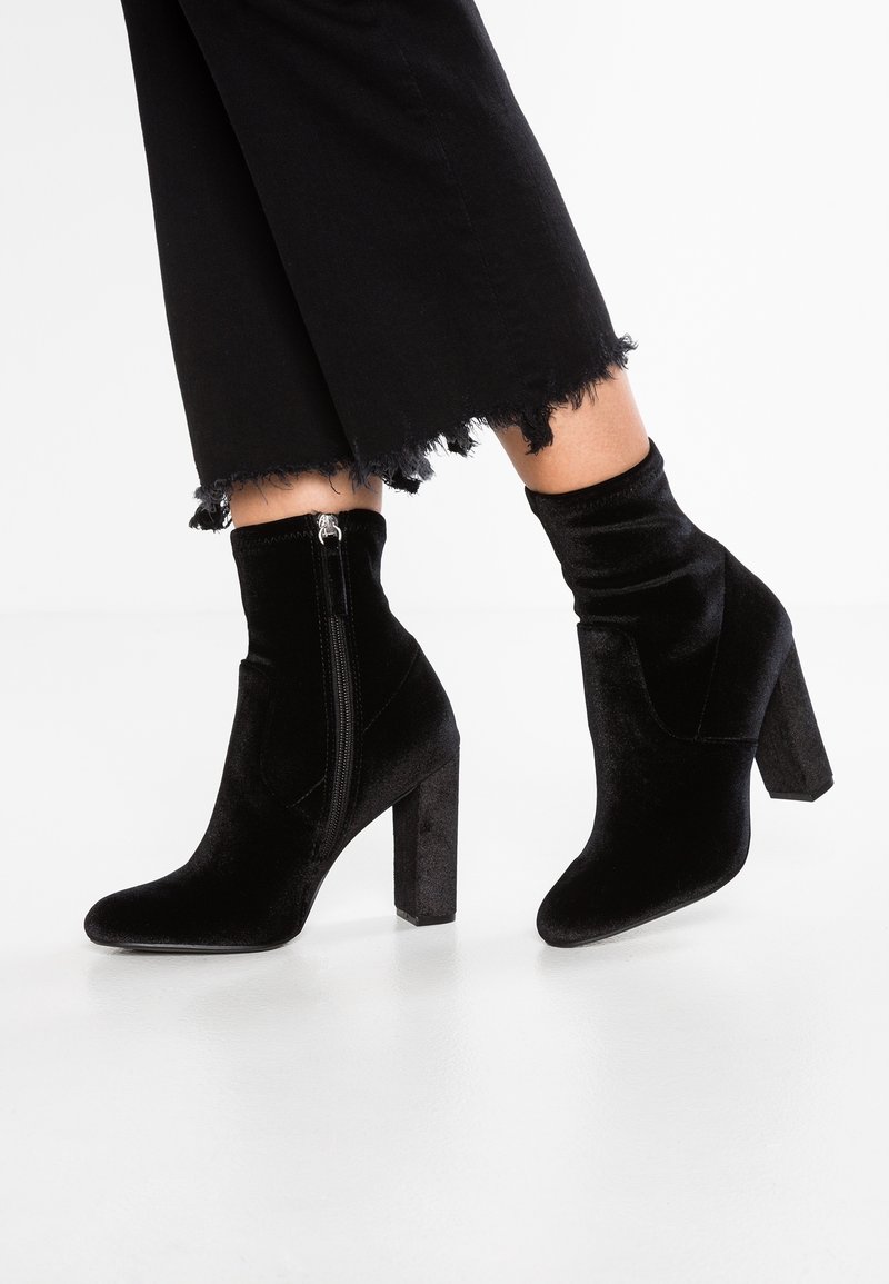 Femme portant des bottines en velours noir à talons carrés et fermetures éclair latérales, associées à un pantalon noir court à ourlet effiloché.
