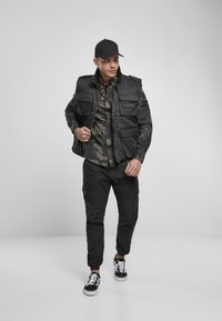 Svart puffervest over en kamuflasje-skjorte med knapper, kombinert med svarte cargo-bukser og svarte joggesko med hvite detaljer, alt satt mot en nøytral bakgrunn.