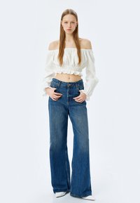 Hvid off-the-shoulder crop top med volang i kanten, parret med højtaljede, vide blå jeans. Modellen står med hænderne i lommen.